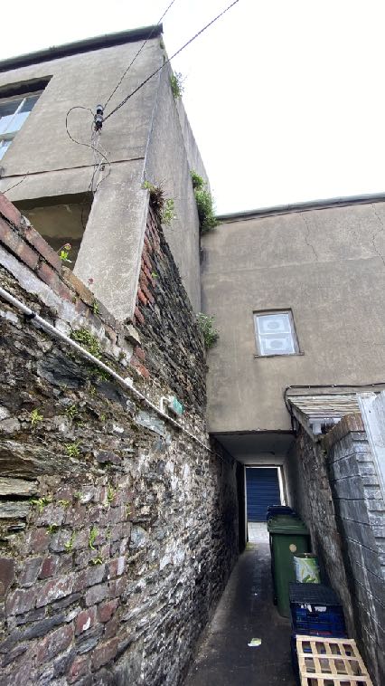 A photograph showing a narrow rear passageway flanked by a weathered stone wall on the left and a rendered building on the right, with waste bins visible.