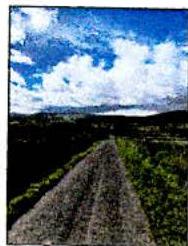 A low-resolution photograph showing a rural dirt track leading towards distant hills under a blue sky with white clouds.