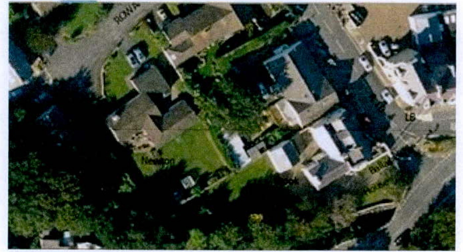 An aerial satellite view showing a residential area with houses, gardens, and roads, likely serving as a location map.