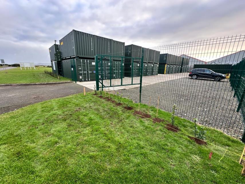 A photograph showing a row of stacked green shipping containers used as self-storage units in an industrial yard, viewed from behind a green fence and grassy verge.