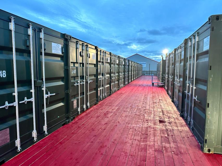 A photograph showing rows of dark green shipping containers arranged on a reddish wooden deck, serving as self-storage units.