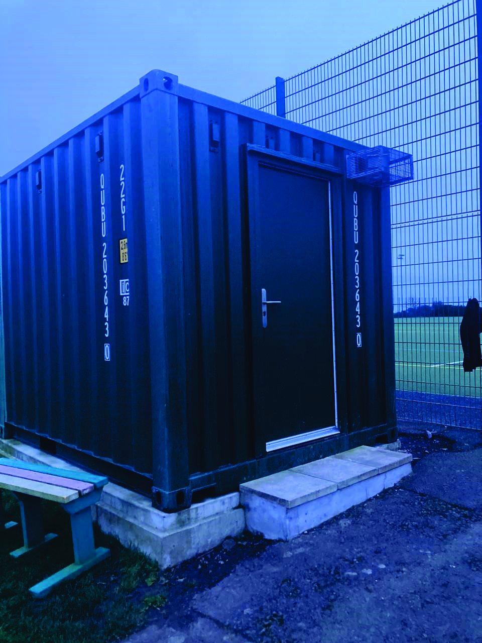 A photograph showing a dark blue shipping container modified with a door, sitting on concrete blocks. It is located next to a tall wire fence bordering a green sports field.