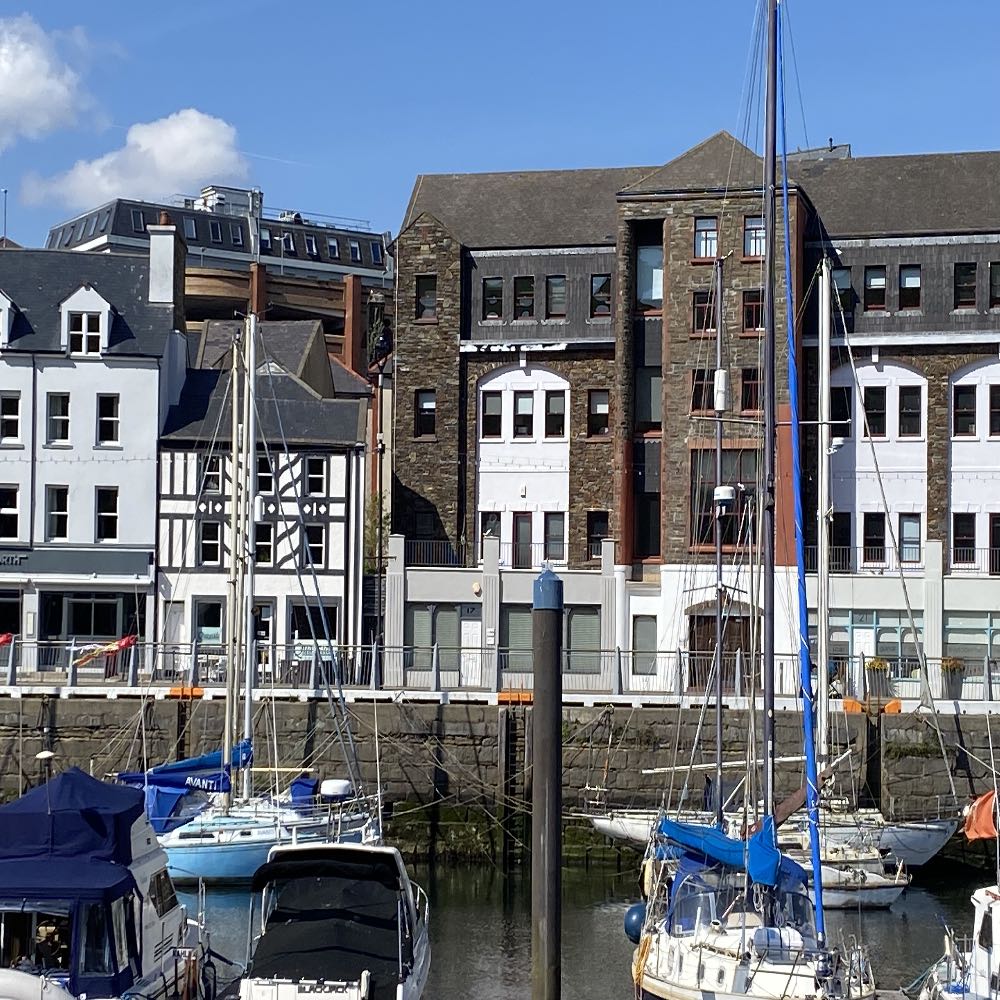 A photograph showing a harbor scene with boats moored in the foreground and a row of multi-story buildings along the quay in the background.