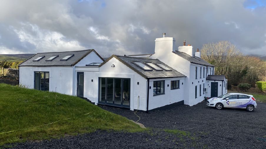 A photograph showing a white, single-story extension or converted agricultural building attached to a farmhouse, featuring a slate roof with skylights and a gravel driveway.