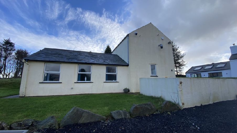 A photograph showing the exterior of a white, single-story building with a slate roof, likely a converted agricultural structure, set in a rural environment with a gravel driveway.