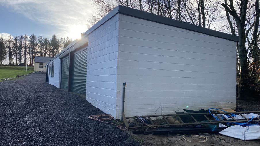 A photograph showing a long, white-painted block building with roller shutter doors, situated on a gravel driveway in a rural setting.