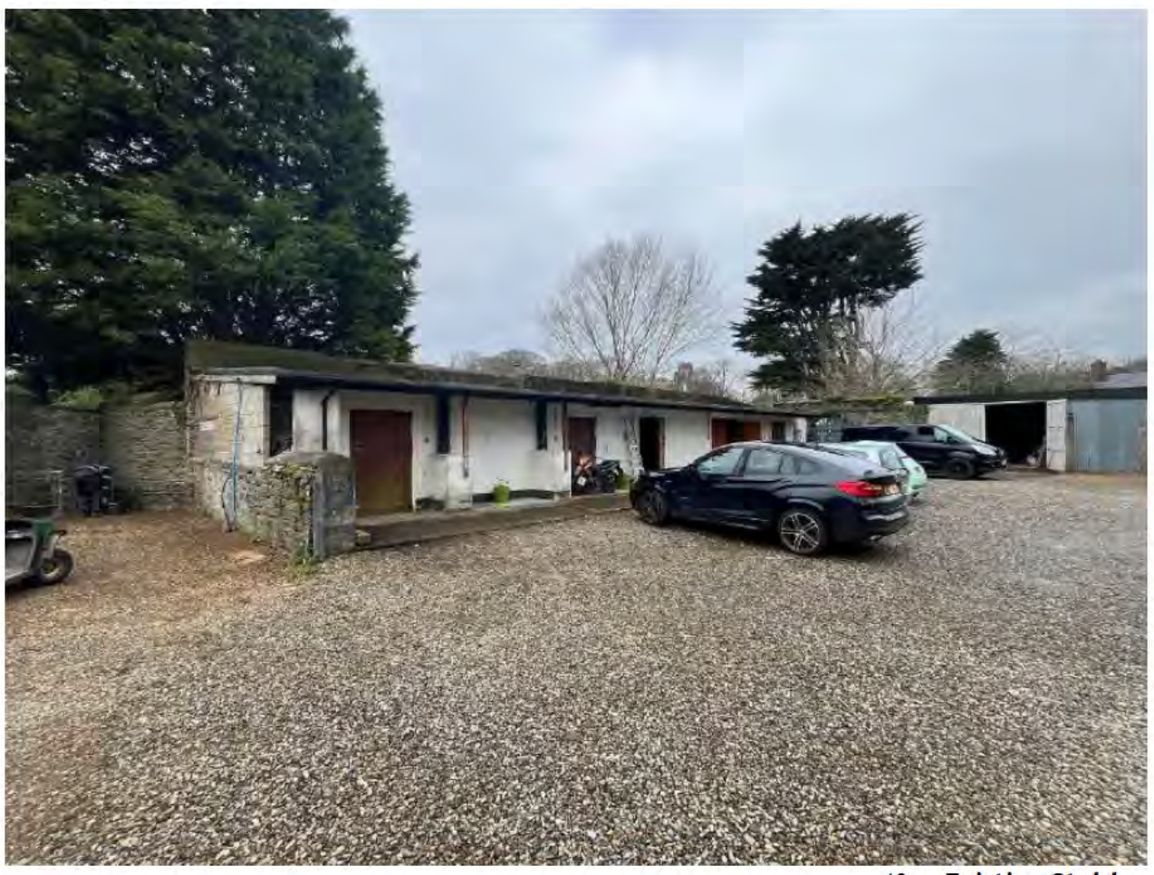 A photograph showing an existing single-story white building with multiple doors, likely garages or small units, adjacent to a gravel parking area with several cars.