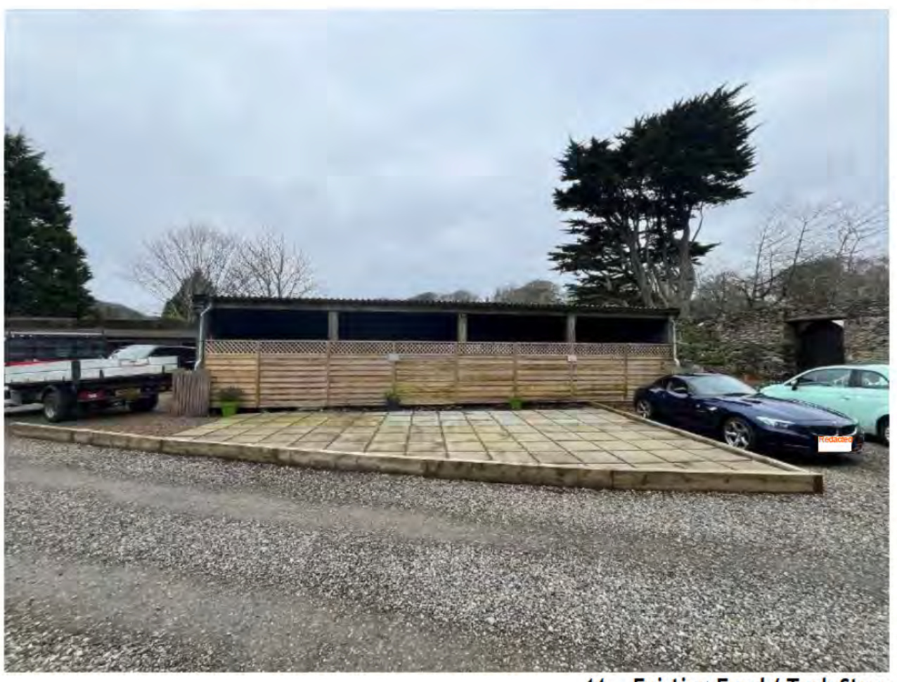 A photograph showing an existing site with a paved parking area, a trailer, and a long wooden structure resembling a stable or covered track with fencing.