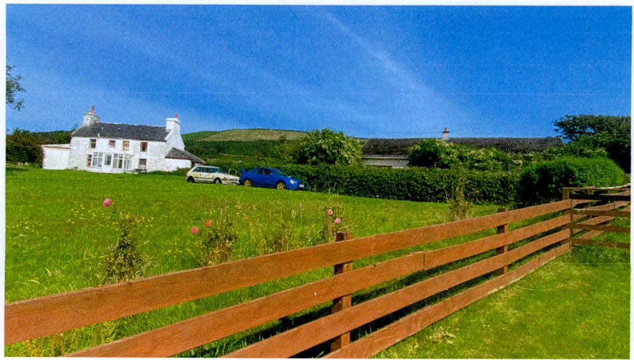A photograph showing a white two-story house situated on a grassy slope with a wooden fence in the foreground and rolling green hills in the background.