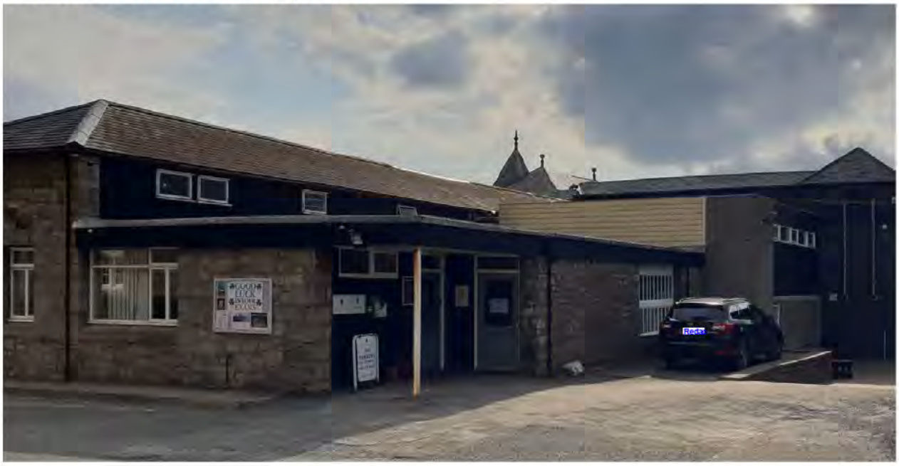 A photograph showing the exterior of a single-story stone building, likely a commercial or community facility, with a car parked in front.