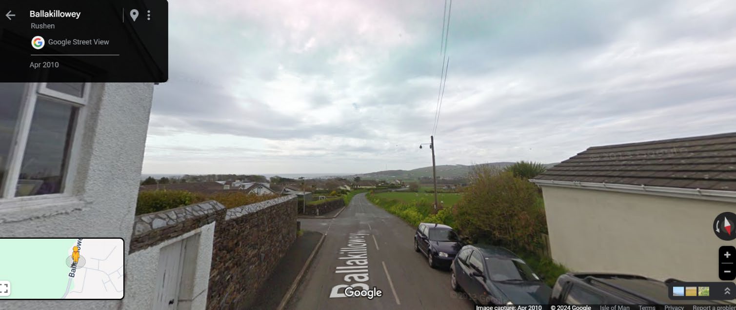 A Google Street View screenshot showing a rural road scene in Ballakilley, Rushen, with houses on either side and a view of the coast in the distance.