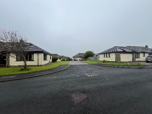 A street-level photograph showing a residential road with detached bungalows on either side under an overcast sky.