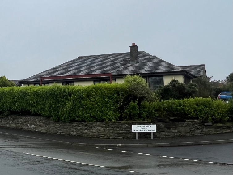 A photograph showing a detached bungalow with a slate roof, situated behind a tall green hedge and stone wall along a roadside.