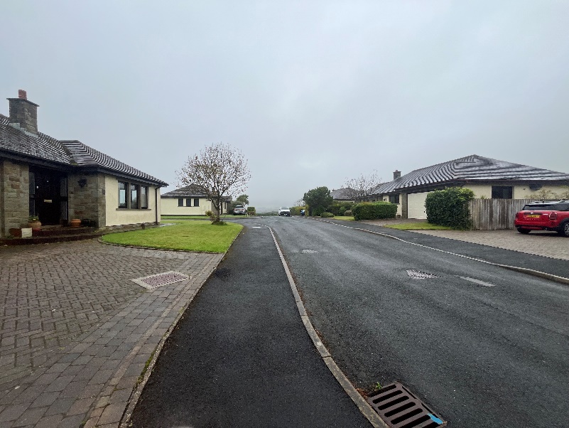 A street-level photograph showing a residential road in Rushen with detached houses featuring stone and rendered facades under an overcast sky.