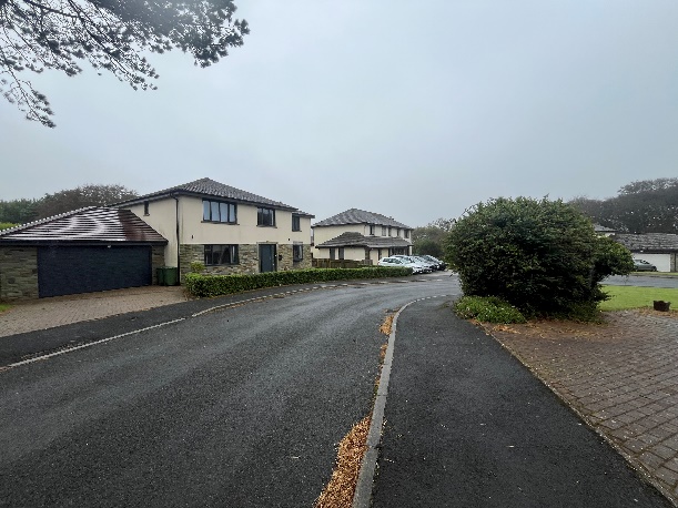 A street-level photograph showing a residential road with detached houses featuring cream render and stone cladding. The image captures the existing property, a garage, and surrounding landscaping.