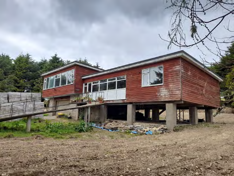 A photograph showing a red wooden bungalow-style house elevated on concrete stilts with a wooden ramp leading to the entrance.