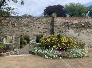 A photograph showing a stone wall with an arched opening, a paved patio area, and flower beds with yellow and white flowers.