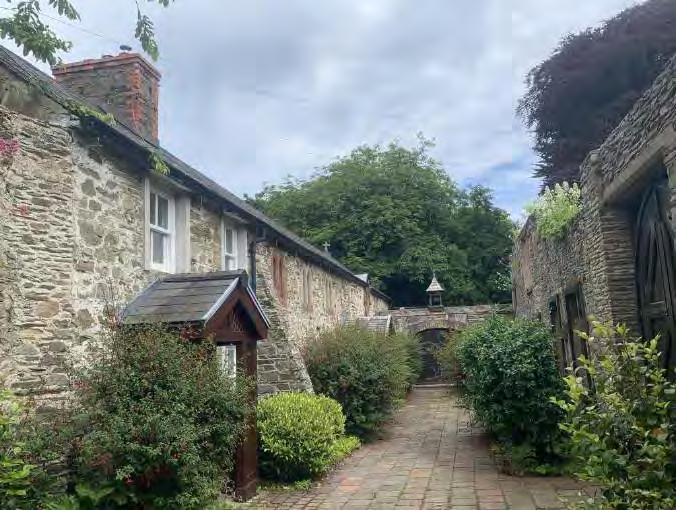 A photograph showing a paved pathway flanked by traditional stone walls and buildings, leading towards a stone archway in a rural setting.