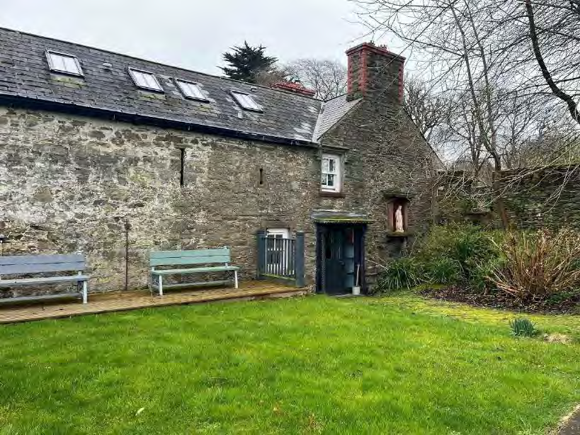 A photograph showing the exterior of a stone building with a slate roof and skylights, set against a green lawn and wooden deck.