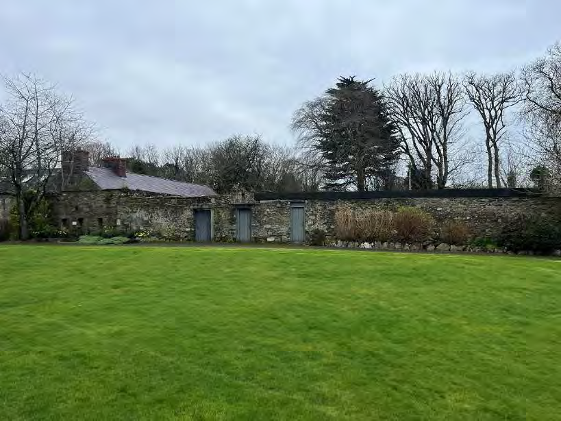 A photograph showing a large grassy area in the foreground with a long stone wall and an old stone building in the background.