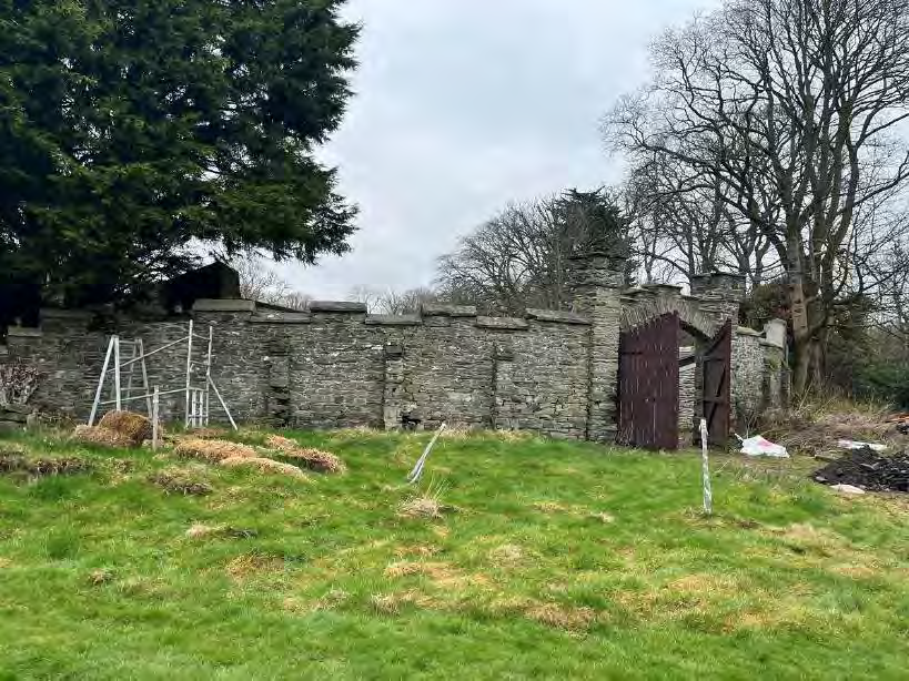 A photograph of a grassy site featuring a long, weathered stone wall and wooden gate in the background. The foreground contains landscaping materials and white marker poles, indicating site preparation.
