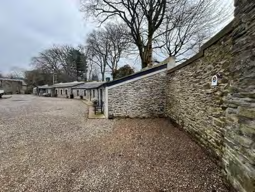 A photograph showing a gravel driveway and a high stone boundary wall, with a row of stone buildings visible in the background under an overcast sky.