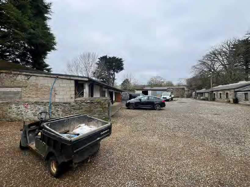 A photograph showing a gravel yard with existing stone agricultural buildings or stables, a utility vehicle in the foreground, and parked cars.
