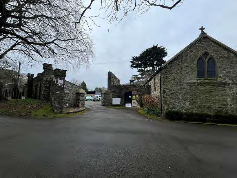 A street-level photograph showing the entrance to a site featuring stone gate piers, a road, and a stone building resembling a chapel with a cross on the roof.