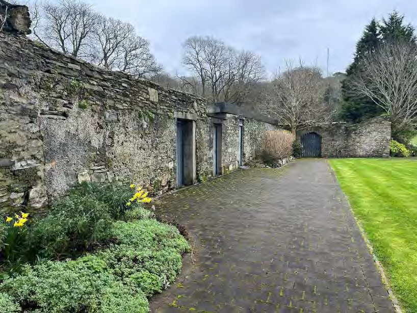 A photograph showing a long, weathered stone wall with multiple doorways alongside a paved driveway and manicured lawn.