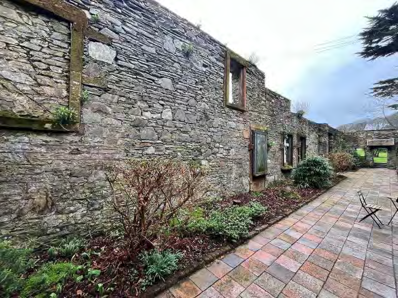A photograph showing the exterior of a long, weathered stone wall with empty window openings, situated next to a paved patio area with landscaping.