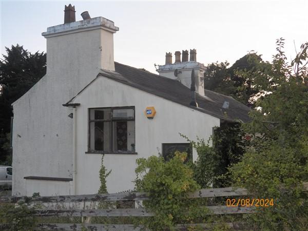 A photograph of a white, single-story detached building with prominent chimney stacks and overgrown vegetation in the foreground.
