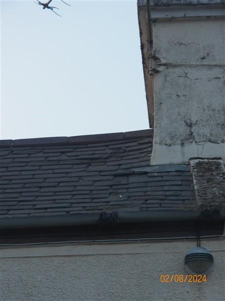 A close-up photograph of a building corner showing a slate roof, white rendered wall, and gutter, with a bird flying in the sky above.