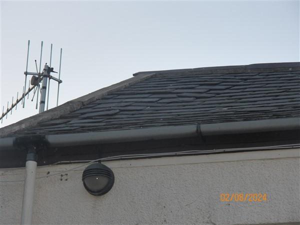 A close-up photograph of a building's roofline featuring slate tiles, a gutter, downpipe, and a television aerial against a grey sky.