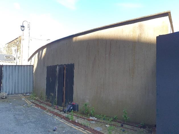 A photograph showing the exterior side of a large, curved concrete building with dark openings, situated next to a corrugated metal fence.