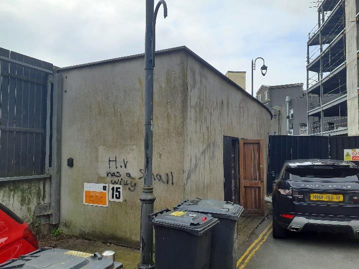 A street-level photograph showing a small, weathered concrete building with graffiti, situated next to a construction site with parked cars and bins in the foreground.