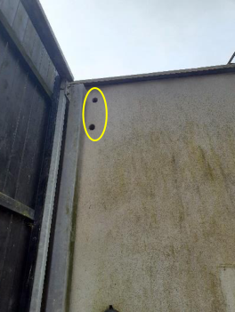 A close-up photograph showing the corner of a rendered wall next to a wooden fence. A yellow oval highlights two small holes in the wall, indicating a bat or bird survey inspection.