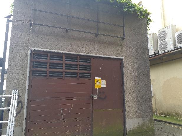 A photograph showing the exterior of a building featuring a large brown metal door with ventilation grilles and air conditioning units mounted nearby.