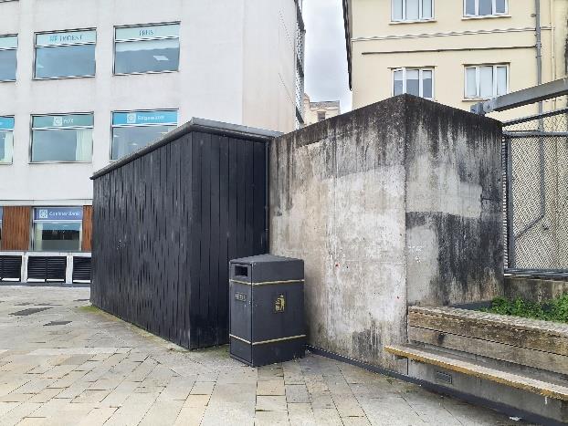A photograph showing a dark wooden outbuilding or bin store situated next to a stained concrete boundary wall and steps. Commercial buildings with large windows are visible in the background.