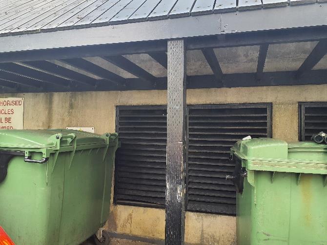 A photograph showing the exterior wall of a building featuring black louvered vents and large green waste bins situated underneath a metal roof structure.