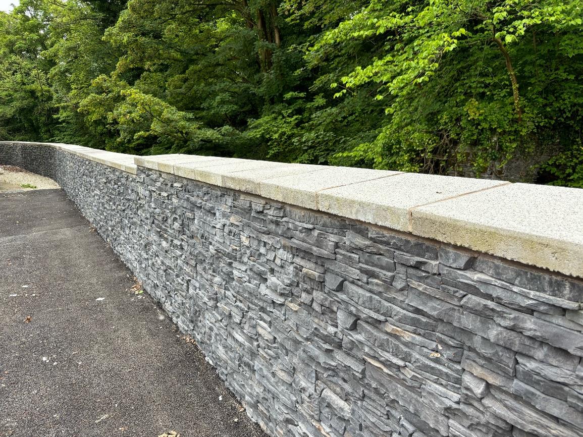 A photograph showing a long, curved stone retaining wall with a light-colored coping stone, running alongside a paved path with dense green trees in the background.