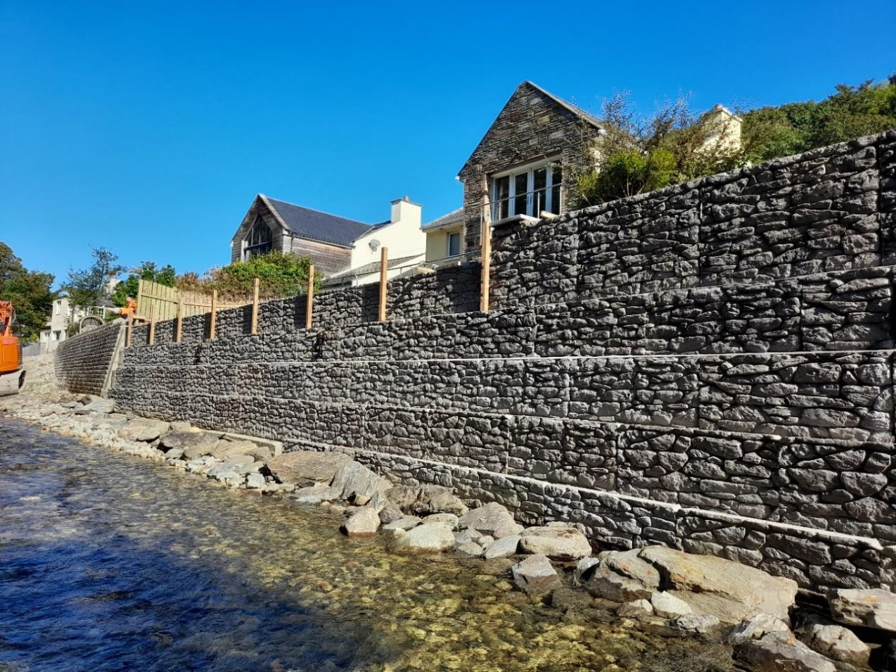 A photograph showing a large, tiered stone retaining wall constructed along a waterfront, with residential buildings visible on the upper level and construction machinery nearby.