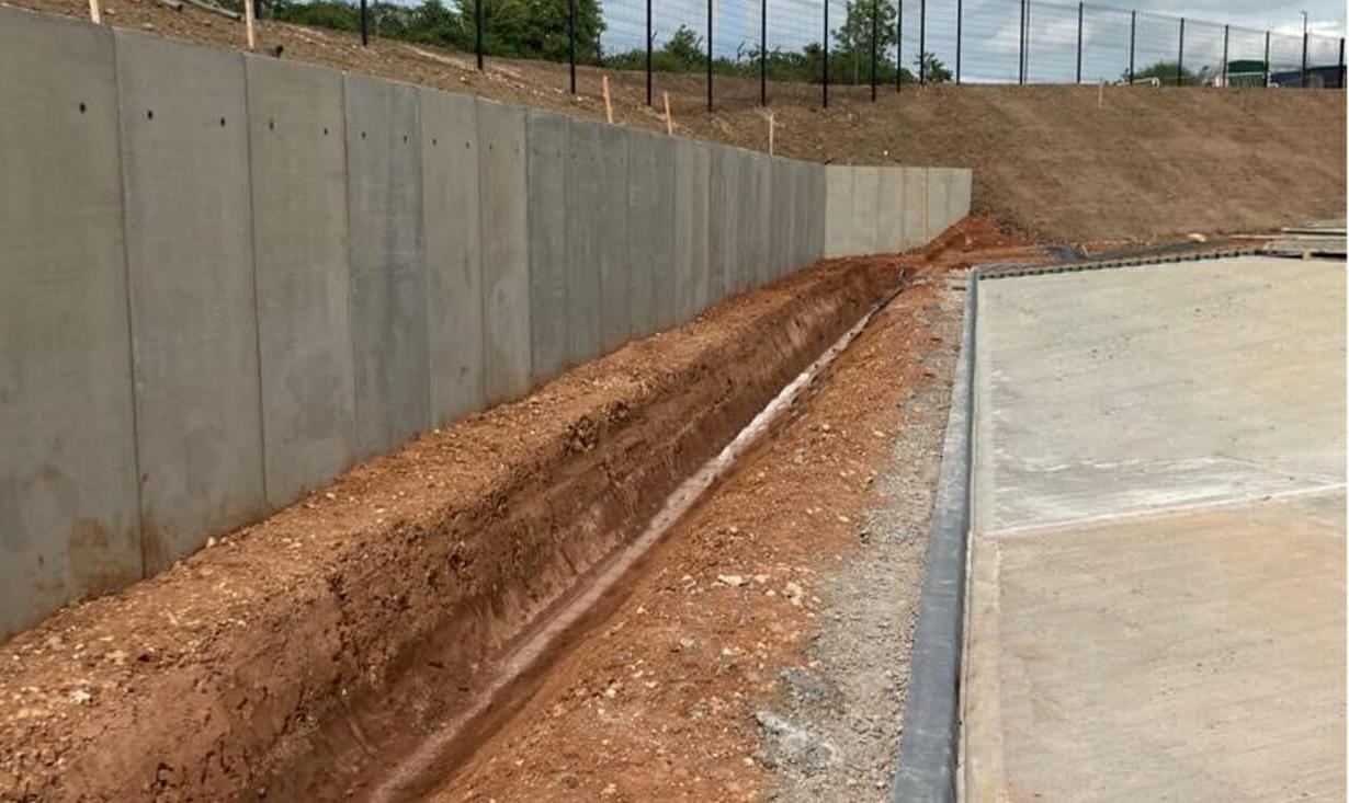 A photograph showing a newly constructed concrete retaining wall with a drainage trench at its base, adjacent to a paved area and an earth bank.