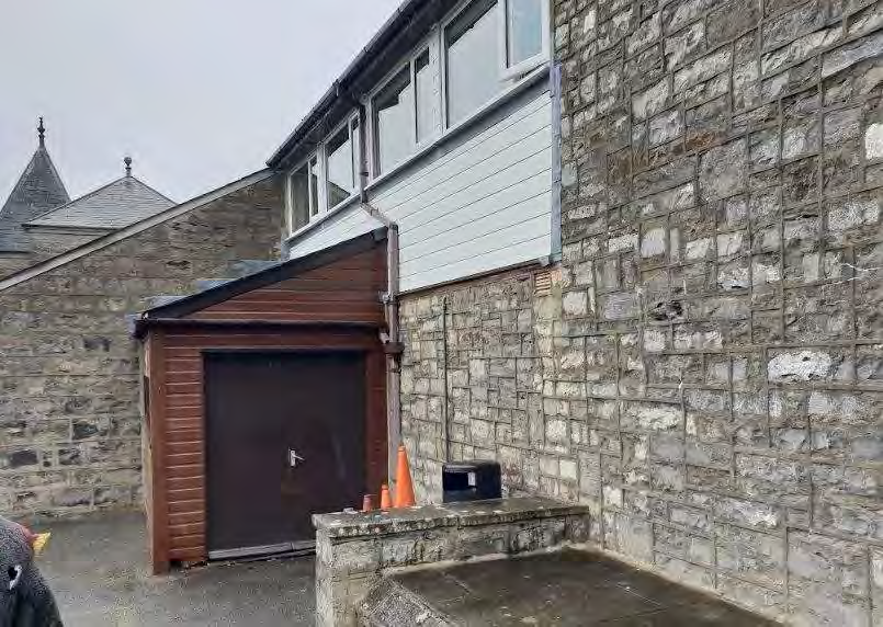 A photograph showing the side elevation of a stone building featuring a wooden garage or porch extension and a low stone boundary wall.