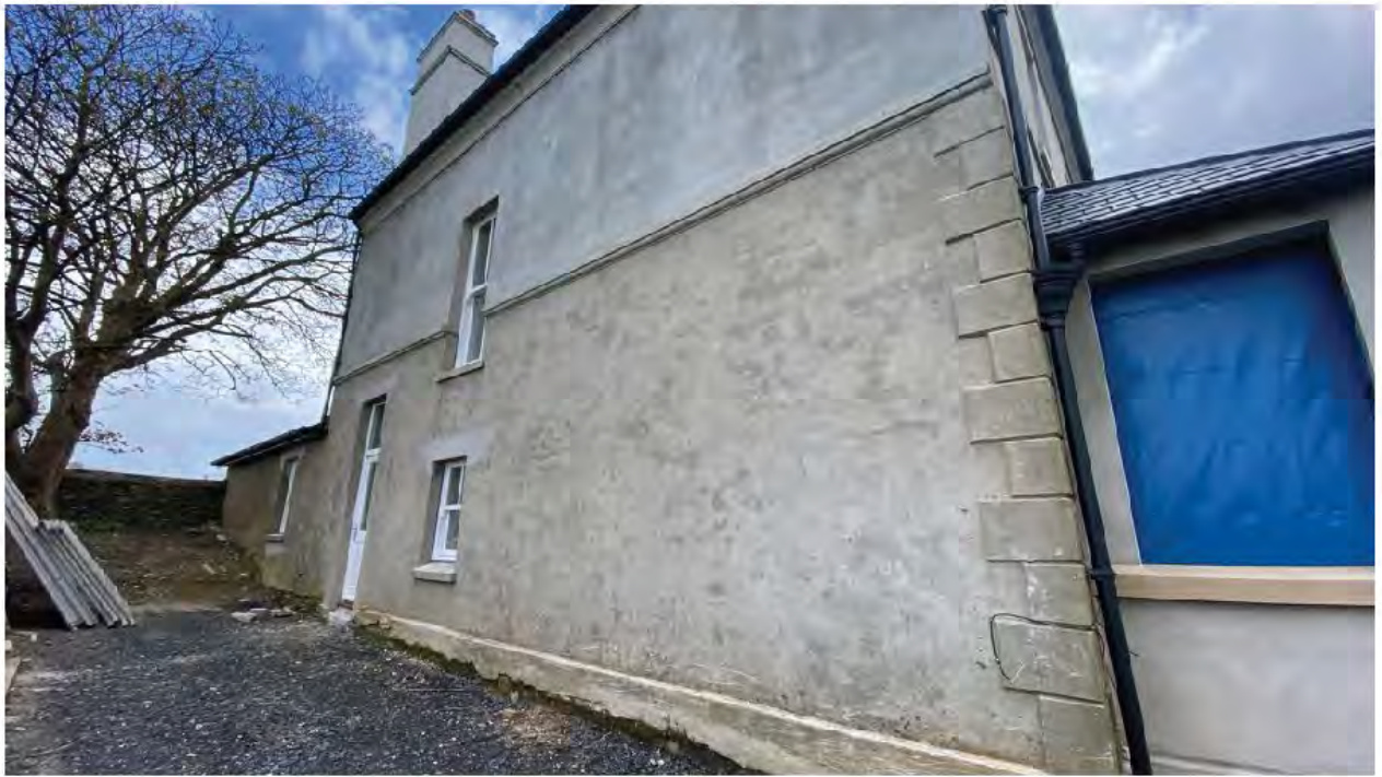 A photograph showing the side elevation of a two-story rendered house with a lower single-story extension on the right featuring a window covered in blue sheeting.