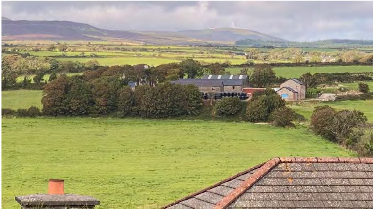 A rural landscape photograph showing green fields and rolling hills in the background, with stone buildings including a long barn-like structure and a smaller outbuilding in the mid-ground.