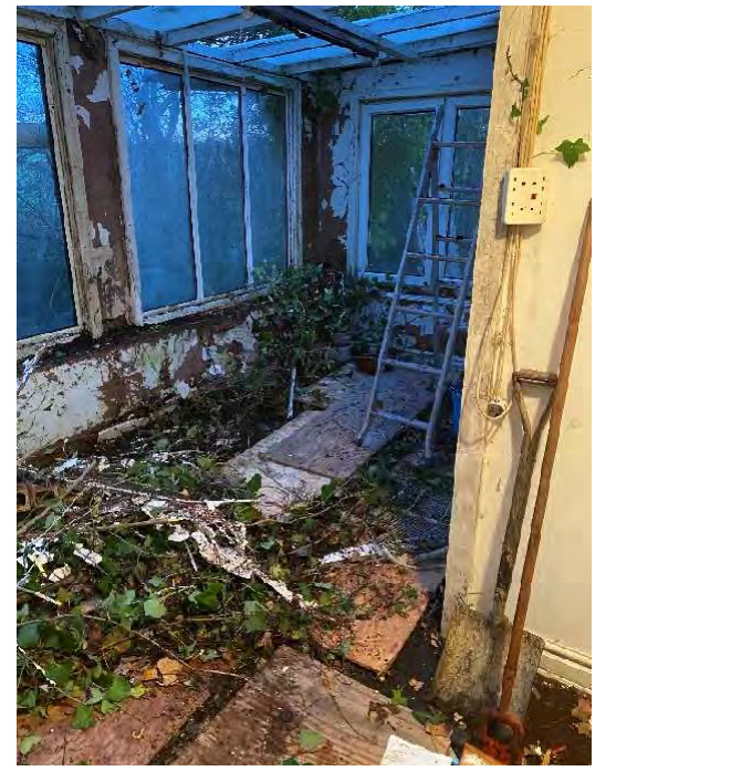 Interior photograph of a dilapidated conservatory or sunroom with peeling paint, debris on the floor, and a ladder leaning against the wall.