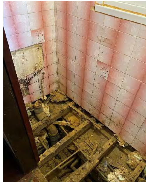 Interior photograph showing a room with pink tiled walls and a floor stripped to expose wooden joists and drainage pipes.
