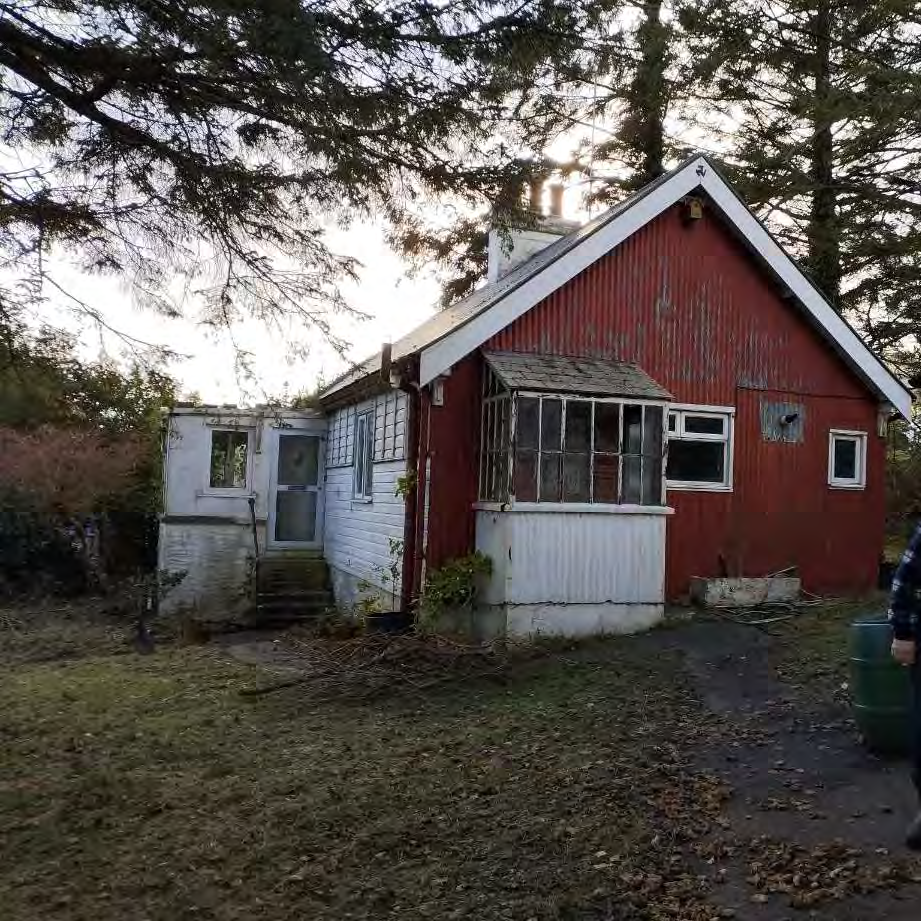 A photograph of an existing single-story dwelling featuring red corrugated cladding and a white side extension, situated in a rural, tree-filled setting.