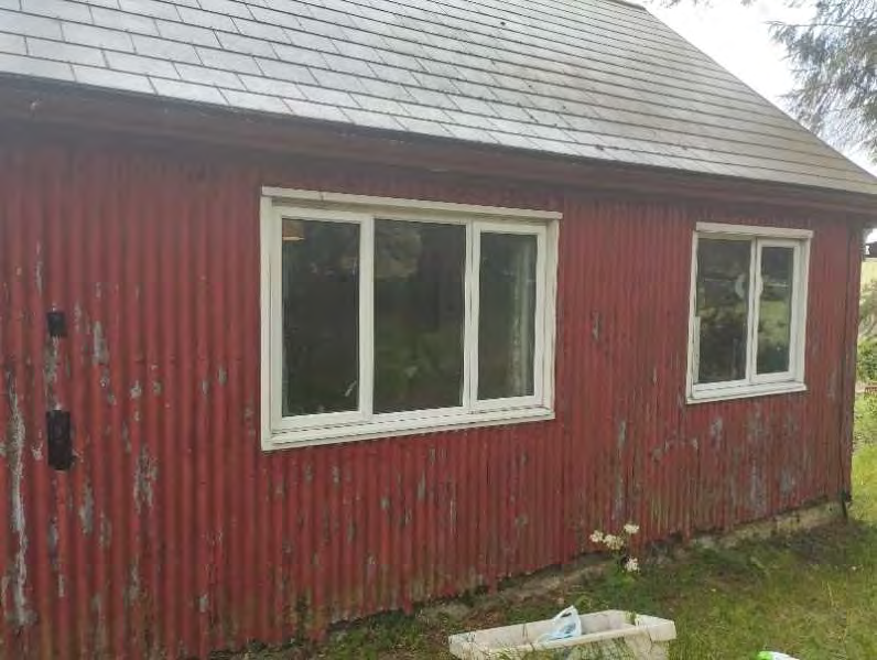 A photograph showing the side elevation of a single-story building with red corrugated cladding and white-framed windows, likely an existing garage or outbuilding.
