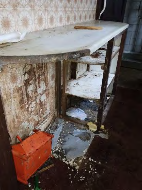 Interior photograph of a dilapidated room showing a damaged worktop, rotting shelving, and debris on the floor.